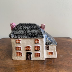 May include: A ceramic house ornament with a white facade, black tiled roof, and pink chimneys. The windows have orange frames. The front door is brown. The ornament is on a wooden surface.