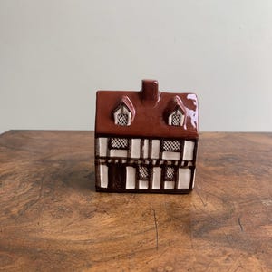 May include: A small ceramic house ornament with a brown roof and white and brown walls. The house features two dormer windows and a chimney. It is placed on a wooden surface.