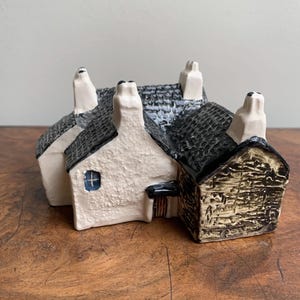May include: A ceramic model of a village scene with three attached houses. The houses have white, textured walls, black tiled roofs, and white chimneys. One house has a small blue window. The houses are on a wooden surface.