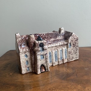 May include: A ceramic miniature church with a brown roof and beige walls. The building features arched windows with blue accents, a clock tower, and a small arched doorway. The design is detailed and textured.