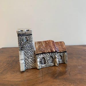 May include: A ceramic church ornament with a textured stone effect. The tower is grey with a white top, and the roof is brown. The arched windows are visible. The ornament is on a wooden surface.