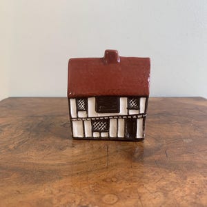 May include: A miniature ceramic house with a red roof, white walls, and black trim. The house features small windows with a crosshatch design and a dark brown door. The overall design is reminiscent of a traditional cottage.