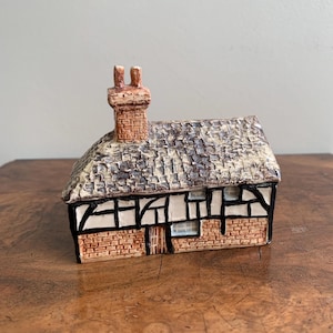 May include: A detailed ceramic miniature cottage with a textured roof, brickwork, and a chimney. The cottage has black and white timber framing and a small door. The design is inspired by traditional English architecture.