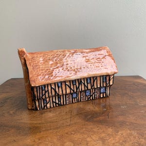 May include: A ceramic house ornament with a brown textured roof and detailed facade. The house features black and blue accents, with a rustic, vintage aesthetic. The ornament is displayed on a wooden surface.