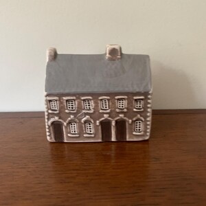 May include: A small ceramic house figurine featuring a grey gabled roof and a brown facade. The structure displays two dark brown arched doorways on the ground floor, flanked by multiple white-framed windows across two levels and a central chimney.