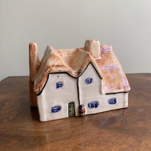 May include: A ceramic cottage ornament with a thatched roof and white walls. The roof is tan and brown, and the walls have blue window details. A small barrel and green door are visible.
