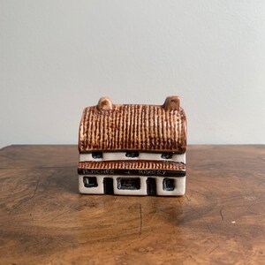 May include: A small ceramic figurine of a building with a brown thatched roof. The building has two storefronts with the words "Butcher + Bakery" written on the front.