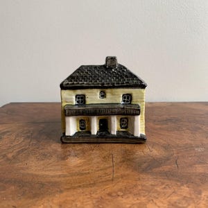 May include: A small ceramic house figurine with a yellow facade, black roof, and white trim. The house has a front porch with three columns and two windows on each side.