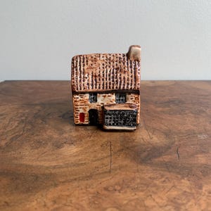 May include: A small ceramic cottage with a thatched roof and a red door. The cottage has two windows and a sign that reads "Post Office".