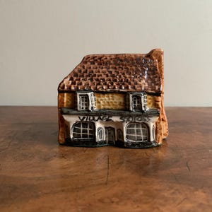 May include: A small ceramic figurine of a village shop with a brown roof, yellow walls, and black windows. The shop has a sign that reads "Village Stores".