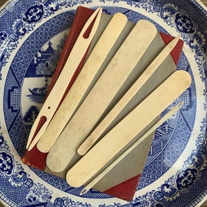 May include: A collection of vintage bone tools, including a needle and various spatulas, arranged on a red and gray book, set on a blue and white patterned plate. The tools vary in size and shape, with a light cream color.
