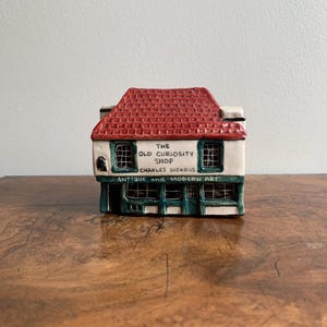 May include: A ceramic miniature replica of a shop with a red roof and green trim. The shop is labelled "The Old Curiosity Shop, Charles Dickens, Antique and Modern Art."