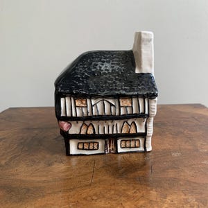 May include: A ceramic house ornament with a black textured roof, white chimney, and a white and black facade. The house features multiple windows with tan and brown details, and a pink heart-shaped accent.