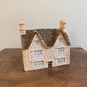 May include: A ceramic house figurine with a textured, off-white facade and a brown, textured roof. The house has two chimneys, small windows, and a brown door. The figurine is set on a wooden surface.