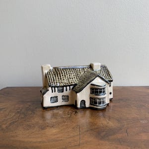 May include: A small ceramic house figurine with a white exterior, brown roof, and black window details. The house has a curved bay window and a chimney.
