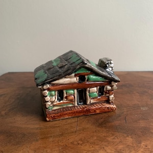 May include: A small ceramic log cabin figurine with a green and brown roof, brown log walls, and a brown porch.
