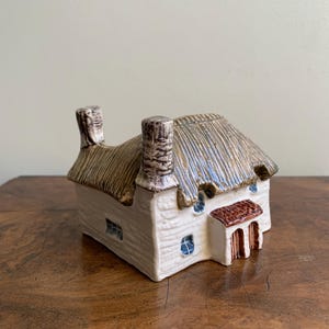 May include: A ceramic cottage ornament featuring a thatched roof and two chimneys. The cottage is painted white with blue window details and a brown door. The roof has a mix of tan and blue tones, giving it a rustic look.