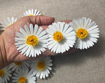 May include: A collection of handcrafted paper daisies featuring white petals and textured yellow centers. Each flower is constructed with layered cardstock to create a three-dimensional appearance, showcasing individual petal shapes and a vibrant yellow core.