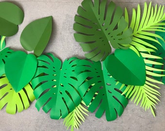 May include: A collection of assorted paper tropical leaves in various shades of green, including large monstera leaves with characteristic cutouts, pointed heart-shaped leaves, and long, slender fern-like fronds arranged in a layered cluster.