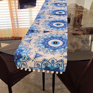Könnte beinhalten: Ein weißer Tischläufer mit einem blauen und weißen Muster, das das böse Auge zeigt. Der Läufer hat einen weißen Pompon-Saum und liegt auf einem Glastisch.