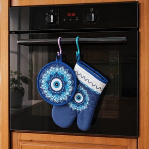 May include: A round blue pot holder and a matching oven mitt featuring a white, light blue, and dark blue evil eye mandala pattern. Both items are suspended from the handle of a black oven by a lavender hook and a turquoise hook respectively.