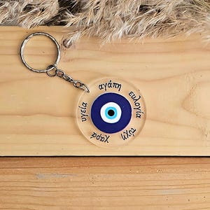 May include: A clear acrylic keychain with a blue evil eye charm and the words "agapi", "ygeia", "xara", "tyxi", "eudoxia" written around the charm.