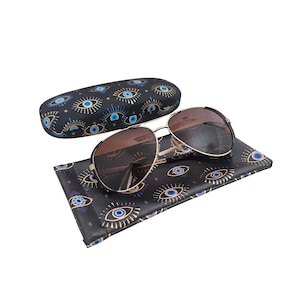 Könnte beinhalten: Goldgerahmte Sonnenbrille mit braunen Gläsern liegt auf einem schwarzen Etui und einem passenden Tuch, beide mit einem sich wiederholenden blauen Augenmuster verziert. Die Sonnenbrille hat eine klassische Aviator-Form.