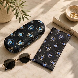 May include: A black hard-shell glasses case and a matching black soft fabric glasses pouch, both featuring a repeating pattern of blue and gold evil eye symbols, alongside a pair of gold-rimmed sunglasses with dark lenses.