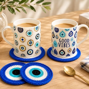 May include: Two white ceramic mugs filled with coffee, one featuring a blue and gold evil eye pattern and the other a blue and gold teardrop pattern with the text GOOD VIBES, resting on blue crocheted coasters with two additional matching coasters.