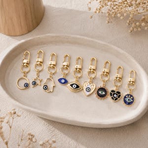 以下が含まれることがあります： ゴールドトーンの金属製キーホルダー9個セット。それぞれにロブスタークラスプとナザールボンジュウ（邪視）モチーフのチャームが付いています。チャームは円形、星型、ハムサの手、ハート、太陽などの形があり、青、白、黒のエナメルで装飾されています。
