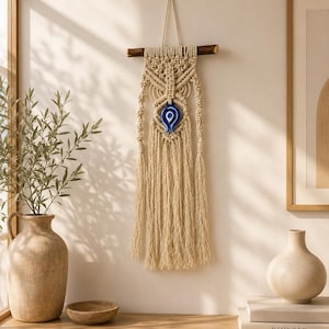 Könnte beinhalten: Ein beigefarbener Makramee-Wandbehang an einem dunklen Holzast mit komplexen Knotenmustern und einem zentralen blau-weißen Glas-Amulett in Form eines Auges. Das Stück endet in langen, losen Fransen, die vertikal vom Ast herabhängen.