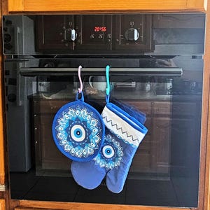 Puede incluir: Dos guantes de horno azules con un diseño de mandala blanco y azul y un amuleto de ojo azul colgado de ganchos sobre un horno negro.