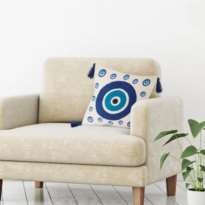 May include: A white throw pillow with a blue and turquoise evil eye design. The pillow has tassels on each corner.