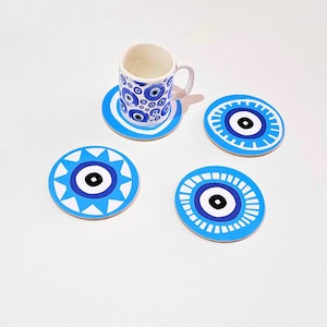 Könnte beinhalten: Ein weißer Keramikbecher und ein passendes Untersetzer-Set mit einem blau-weißen Evil-Eye-Design. Drei zusätzliche Untersetzer mit dem gleichen Design sind um den Becher angeordnet. Die Untersetzer sind rund und aus hellem Material.