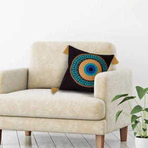 May include: A black throw pillow with a gold and blue evil eye design. The pillow has tassels on each corner.