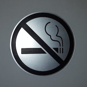 May include: A silver round sign with a black circle and diagonal line through it. The sign has a black image of a cigarette with smoke coming from it. The sign says "No Smoking".
