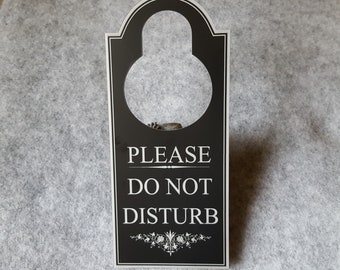 Puede incluir: Un colgador de puerta negro con un círculo blanco y el texto "PLEASE DO NOT DISTURB" en blanco.