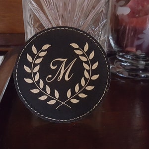 以下が含まれることがあります： 金色のステッチの縁取りと、中央に「M」の文字が入った金色の月桂樹の輪のデザインが施された黒い革製のコースターです。