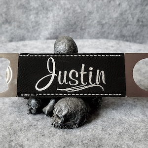 Puede incluir: Un abridor de botellas de cuero negro con un abridor de metal plateado y una inscripción blanca que dice "Justin".