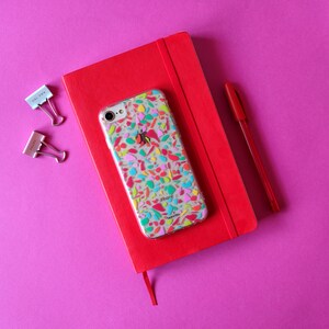 May include: A red notebook with a clear phone case with a colorful mosaic design. The phone case has the word "iPhone" printed on it.
