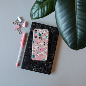 K&ouml;nnte beinhalten: Eine Handyh&uuml;lle mit Terrazzo-Design in Rosa, Grau, T&uuml;rkis und Wei&szlig; liegt auf einem schwarzen Notizbuch mit Punktmuster. Ein rosa Stift und B&uuml;roklammern mit dem Text "STAY SAFE" sind ebenfalls zu sehen.