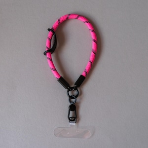 Puede incluir: Un cordón para teléfono de color rosa brillante con un patrón negro. El cordón está hecho de un material de cuerda gruesa y tejida. Tiene herrajes metálicos negros, incluyendo un cierre y un soporte transparente de plástico para el teléfono.