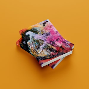 Op de afbeelding: Twee gestapelde notitieboekjes met abstracte, kleurrijke omslagen. De omslagen tonen een mix van roze, paarse, blauwe, groene, gele en zwarte verfspatten en swirls.