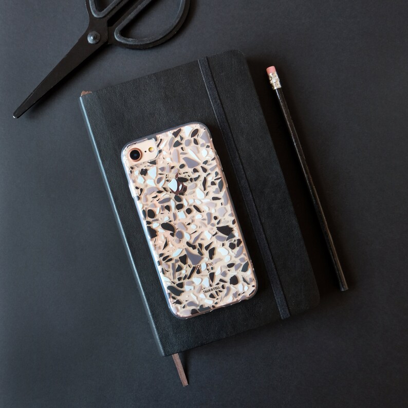 May include: A black notebook with a white and gray patterned phone case on top. The phone case has a white background with a pattern of small, irregular shapes in shades of gray and black.