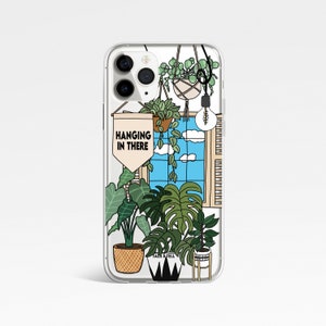 May include: A clear phone case featuring a design of a window with a blue sky and clouds, a lightbulb, and several hanging plants. The text "HANGING IN THERE" is on a banner in the design.
