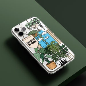 May include: A clear phone case featuring a design of a window with plants hanging from the ceiling. The text "HANGING IN THERE" is on a banner in the design.