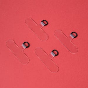 Puede incluir: Cuatro soportes transparentes para teléfono en forma de T con anillos negros, dispuestos sobre un fondo rojo. Estos soportes de plástico transparente están diseñados para adherirse a la parte posterior de un teléfono para un agarre seguro.