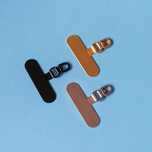 Pode incluir: Três peças de ferragens de metal para bolsas em preto, dourado e ouro rosa, cada uma com uma base retangular e um pequeno fecho preso. As ferragens estão dispostas sobre um fundo azul claro.