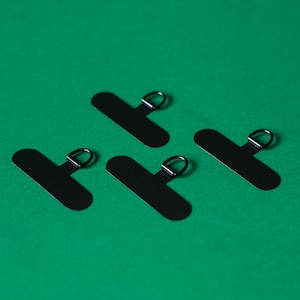 May include: Four black metal hanging tabs with a small metal loop on each. The tabs are on a green background.