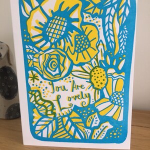 May include: A greeting card with a blue border and a yellow and white floral design. The text "You Are Lovely" is written in the centre of the card.